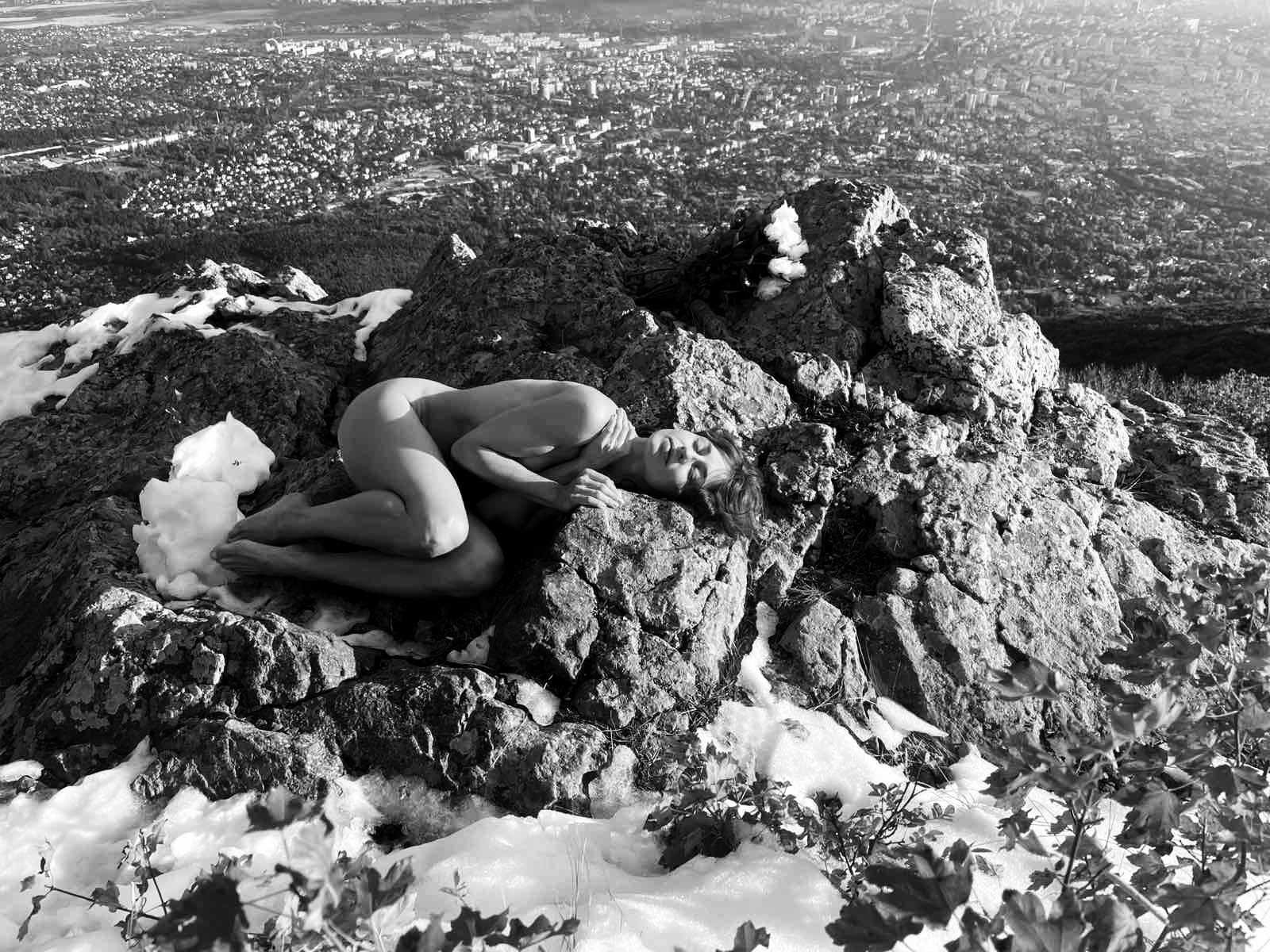 A black and white photograph of a person curled up on rocky terrain, nestled among snow and foliage, with a cityscape visible in the background.